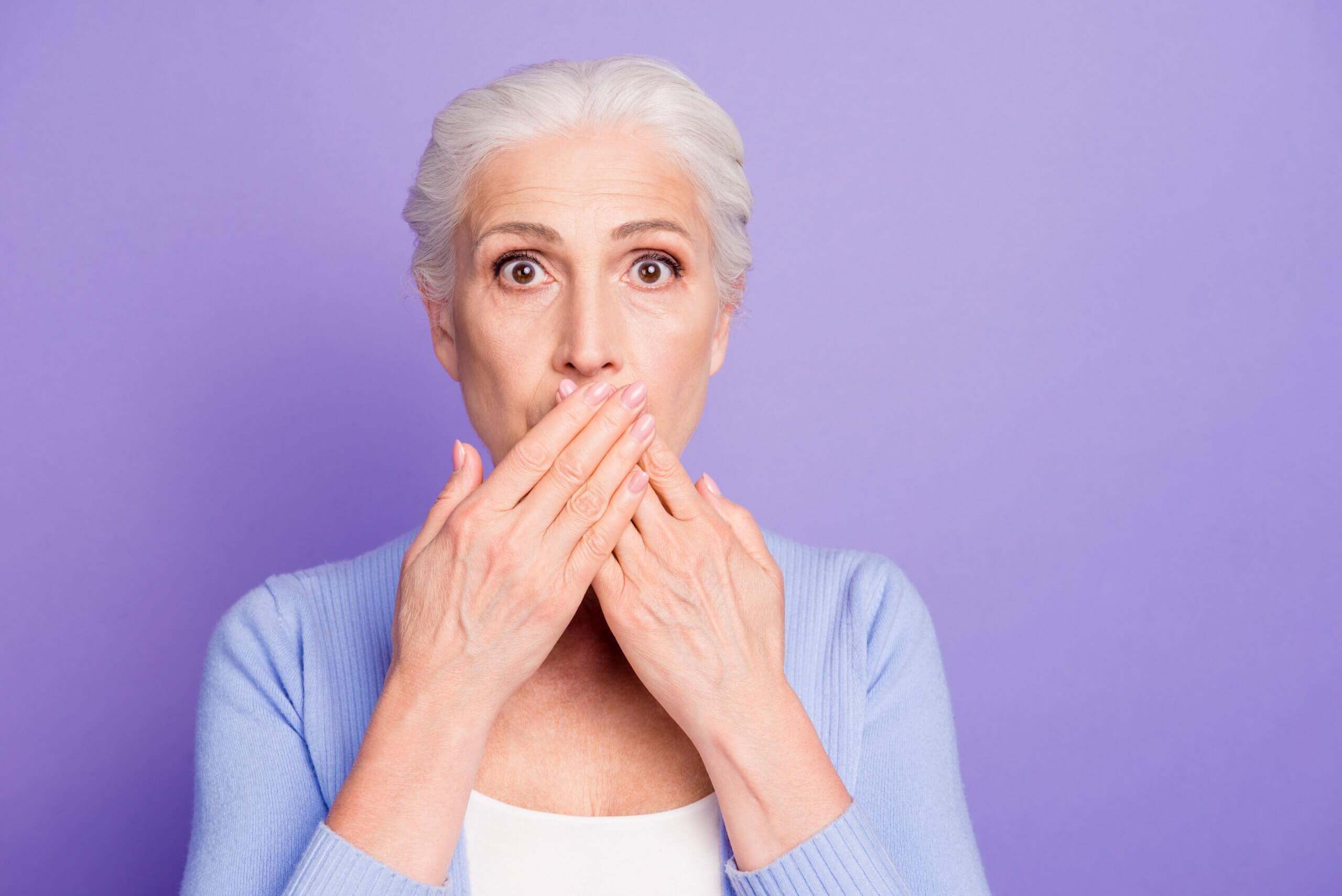 An older woman in front of a violet backdrop with a surprised look in her eyes covering her mouth.